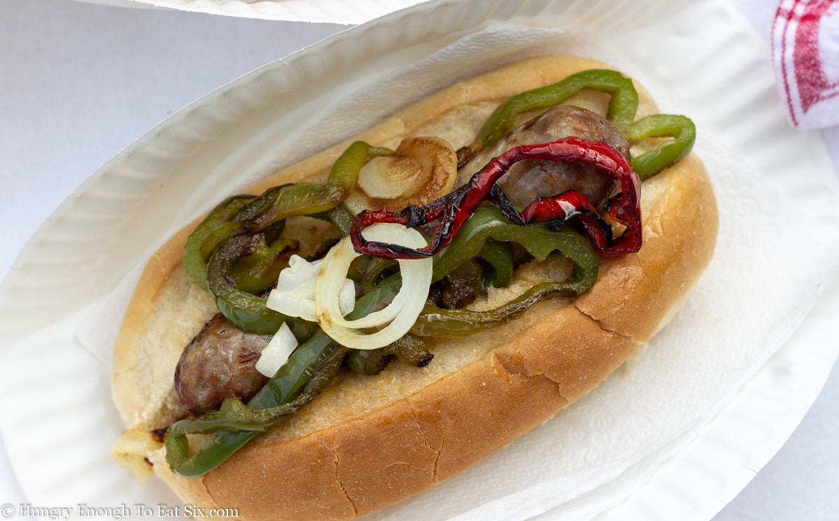 White paper plate holding sub roll with peppers, onions and sausage.