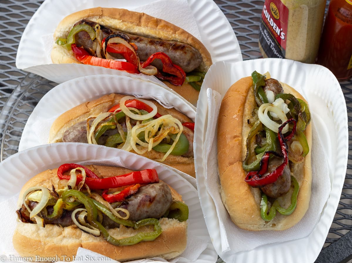 Four sub rolls on paper plates with sausages and peppers and onions.