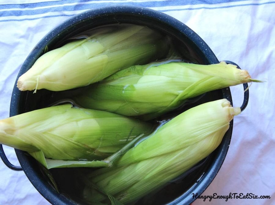 Ears of corn in their husks in a blue pot of water.