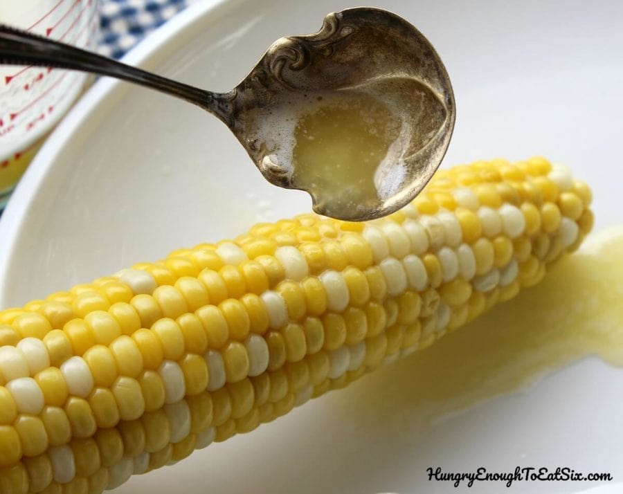 Ear of cooked corn with ladle of butter over it, butter dripping on white plate below.