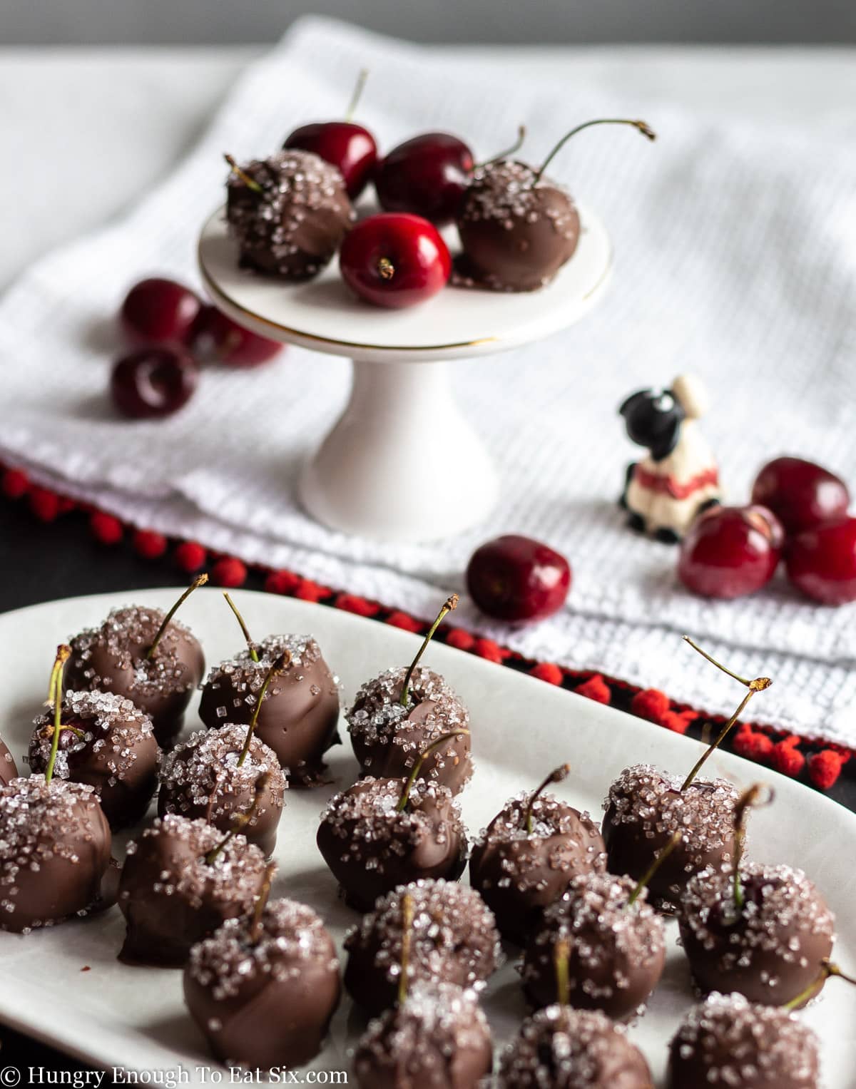 Chocolate and sugar covered cherries on a white platter and pedestal.