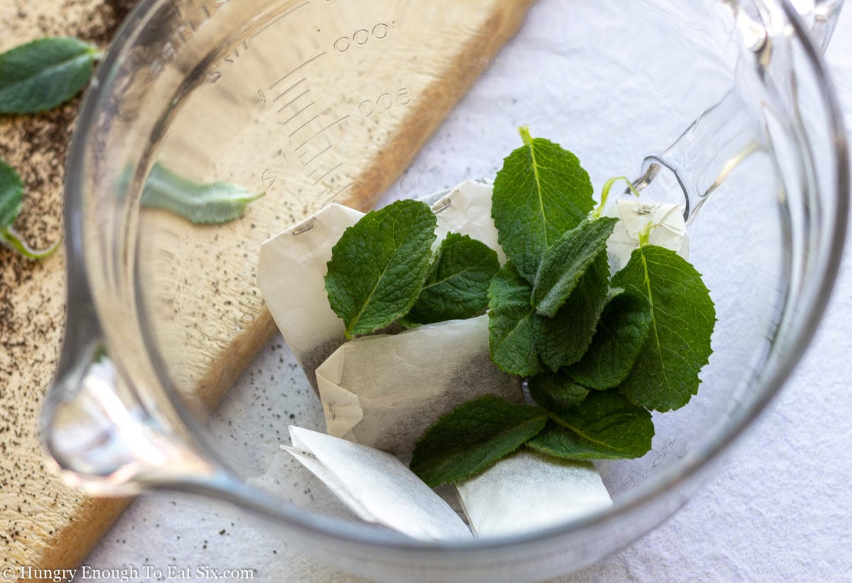 Dry tea bags and fresh spearmint leaves in a large Pyrex pitcher next to a cutting board.