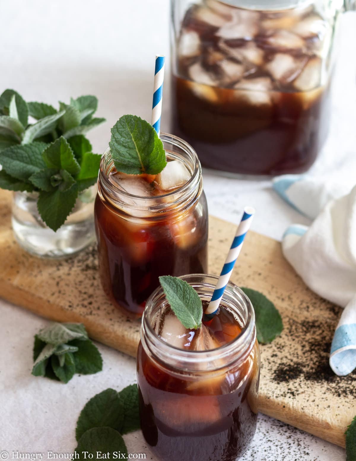 Pitcher and mason jars of dark iced tea with ice cubes and mint leaves.
