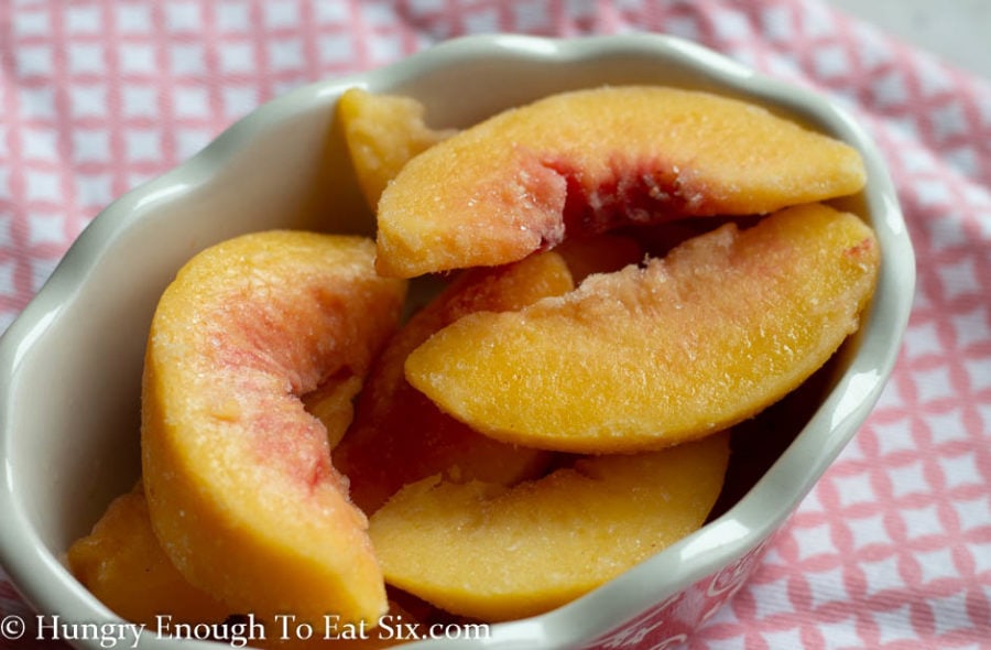 Sliced frozen peaches in a white-rimmed dish on a pink patterned cloth.