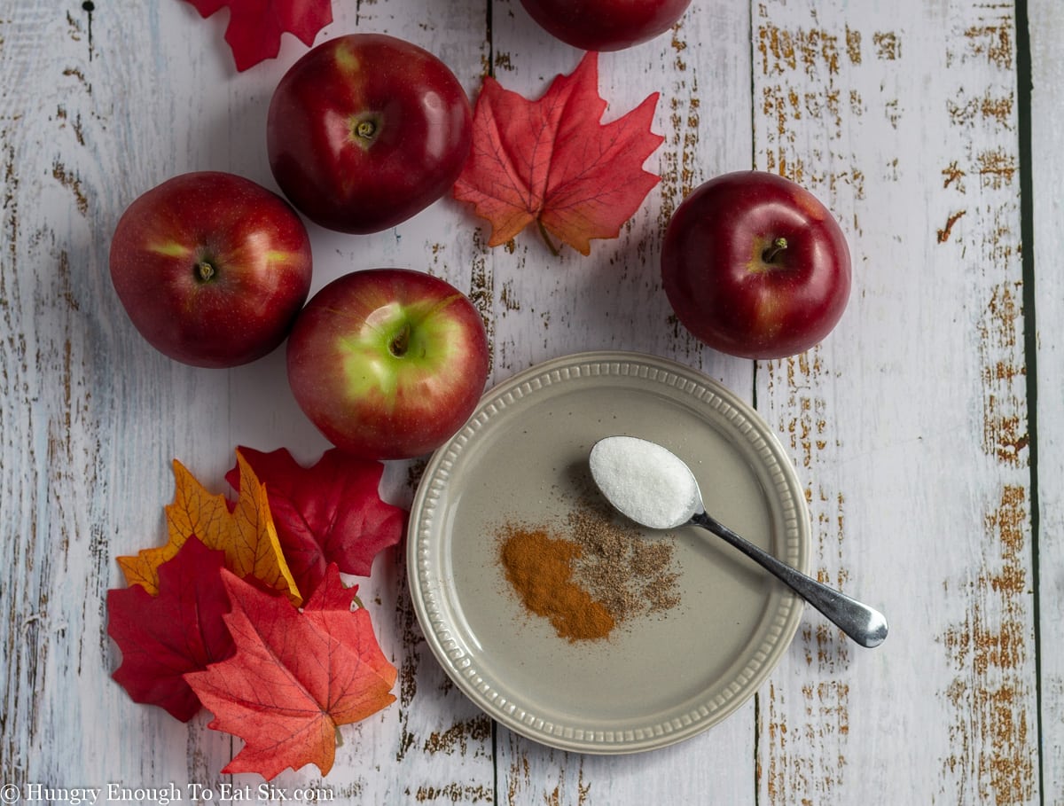 Small plate with piles of spices and a spoon of sugar, with red apples and leaves around.