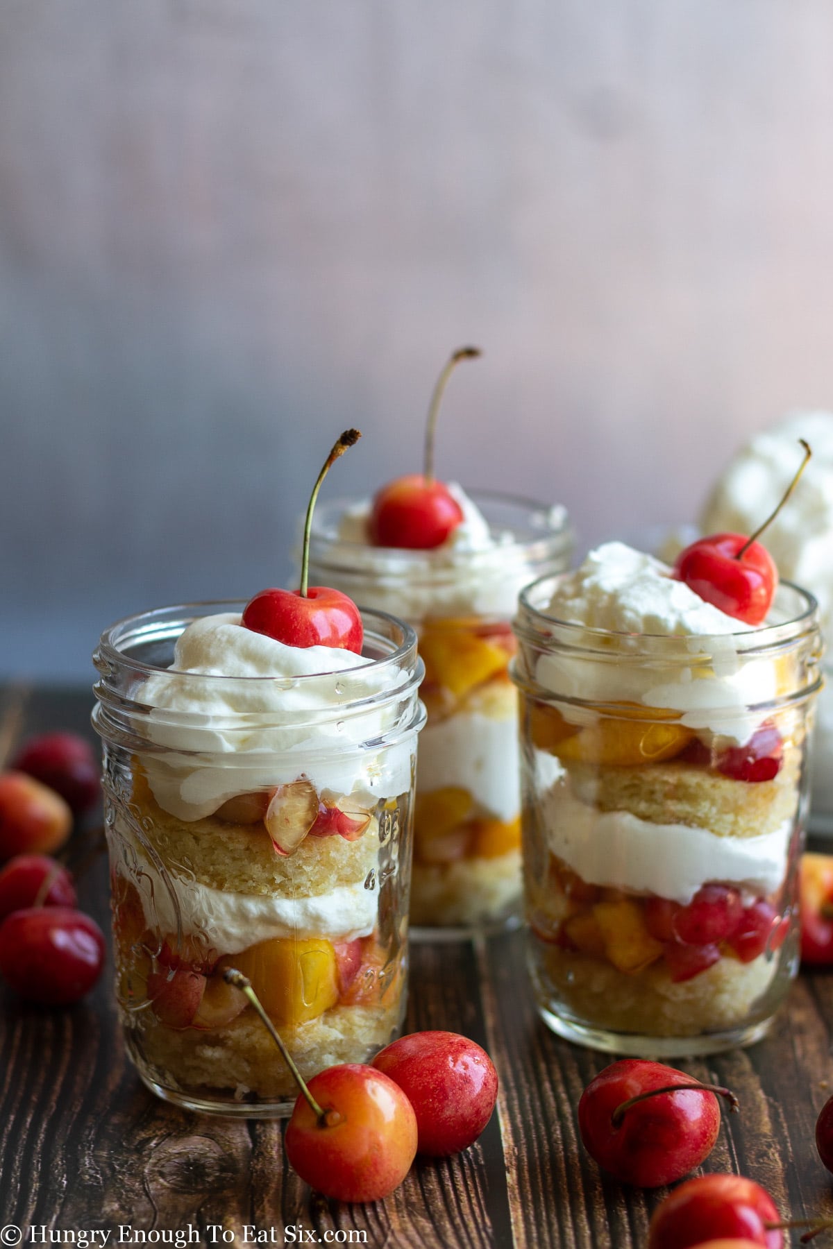 Three cherry nectarine trifles in clear jars with cream, cake and fruit.