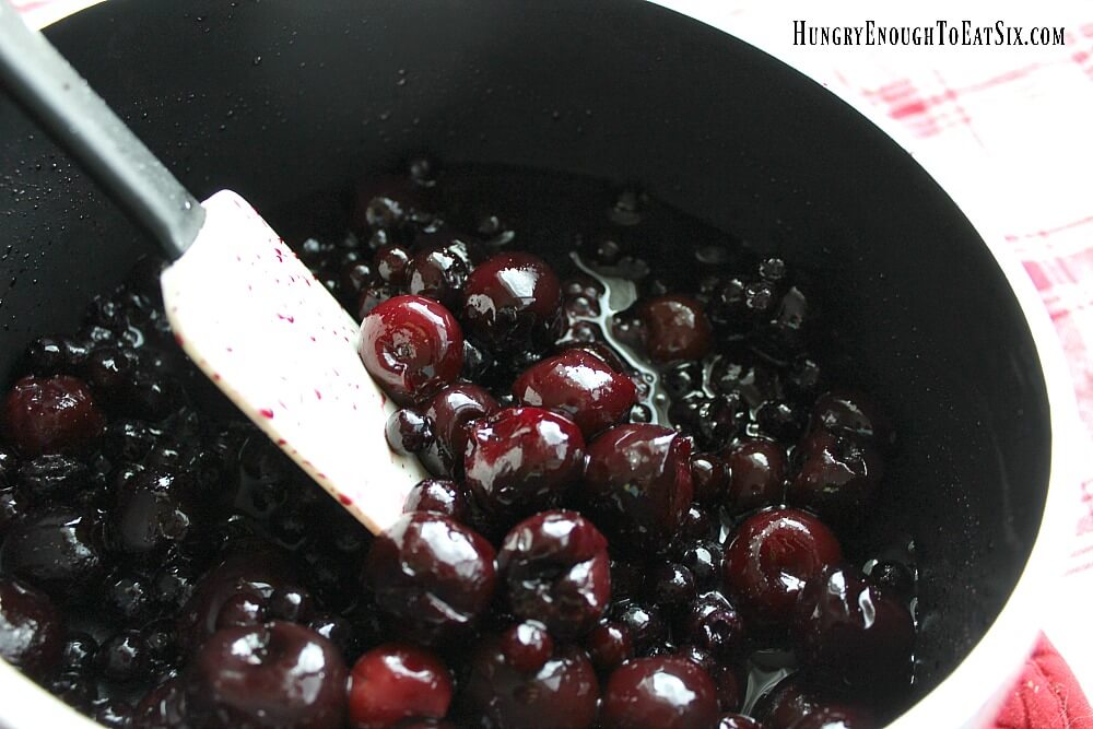 A pan of cherries and blueberries mixed with sugar and cornstarch stirred together, rubber spatula on the side.