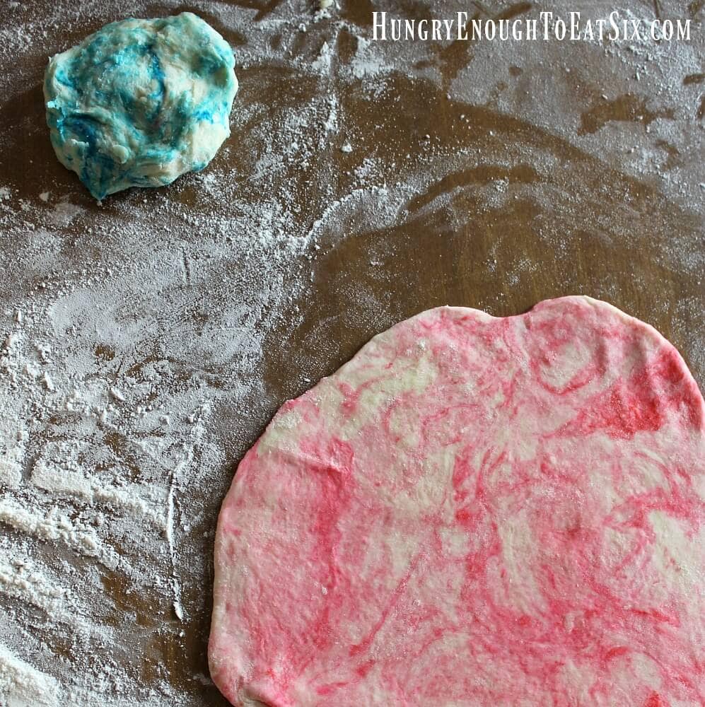 A rolled out circle of red pastry dough on a floury surface, a smaller blue piece of dough in the background.
