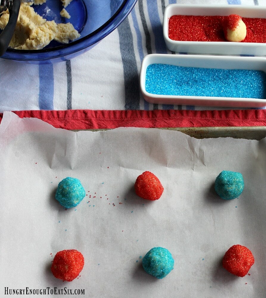 Balls of cookie dough rolled in blue and red sanding sugar on a cookie sheet, dishes of the sugars in the background.