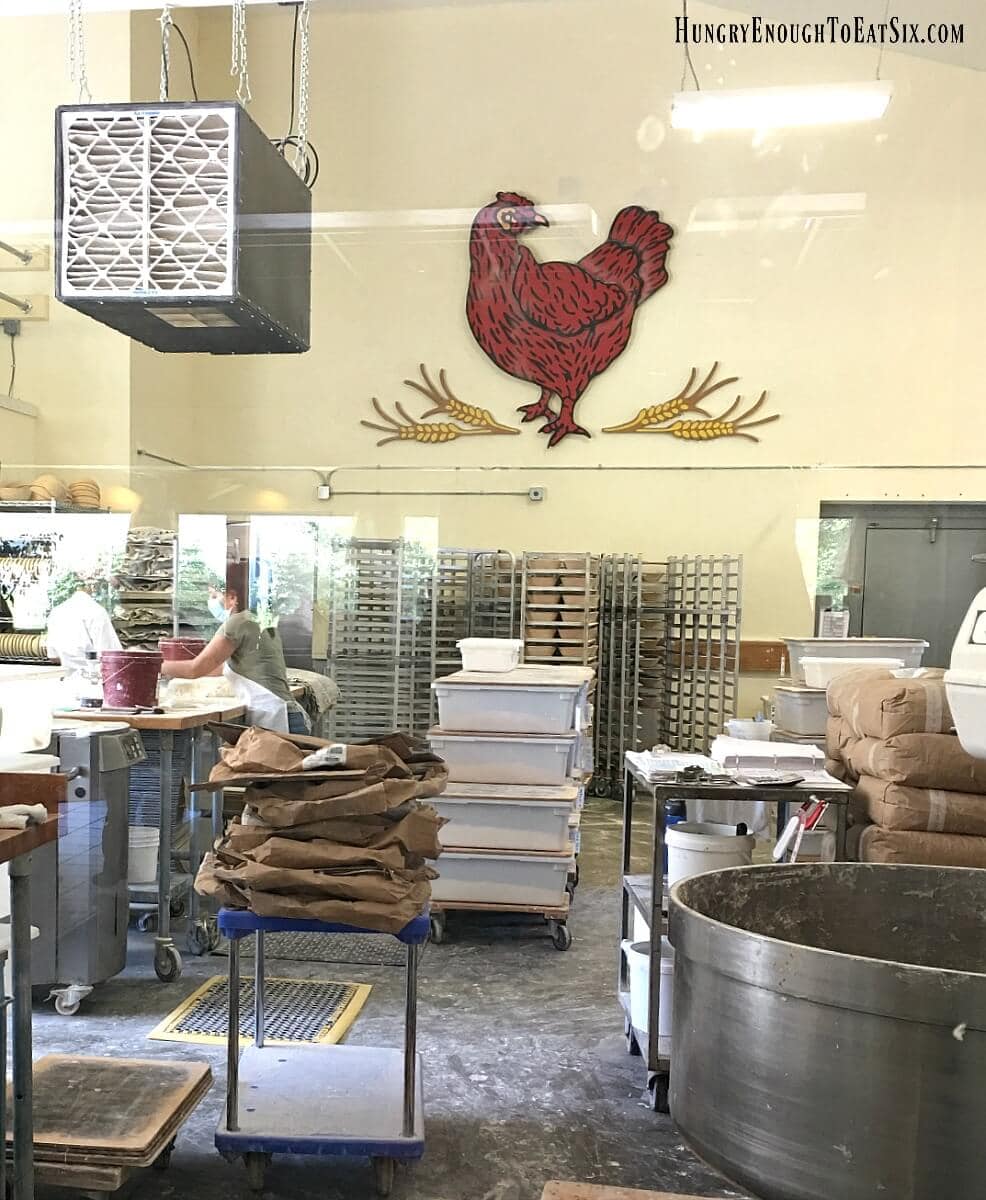 View through a window of bakery operations and a large red hen emblem on the wall. 