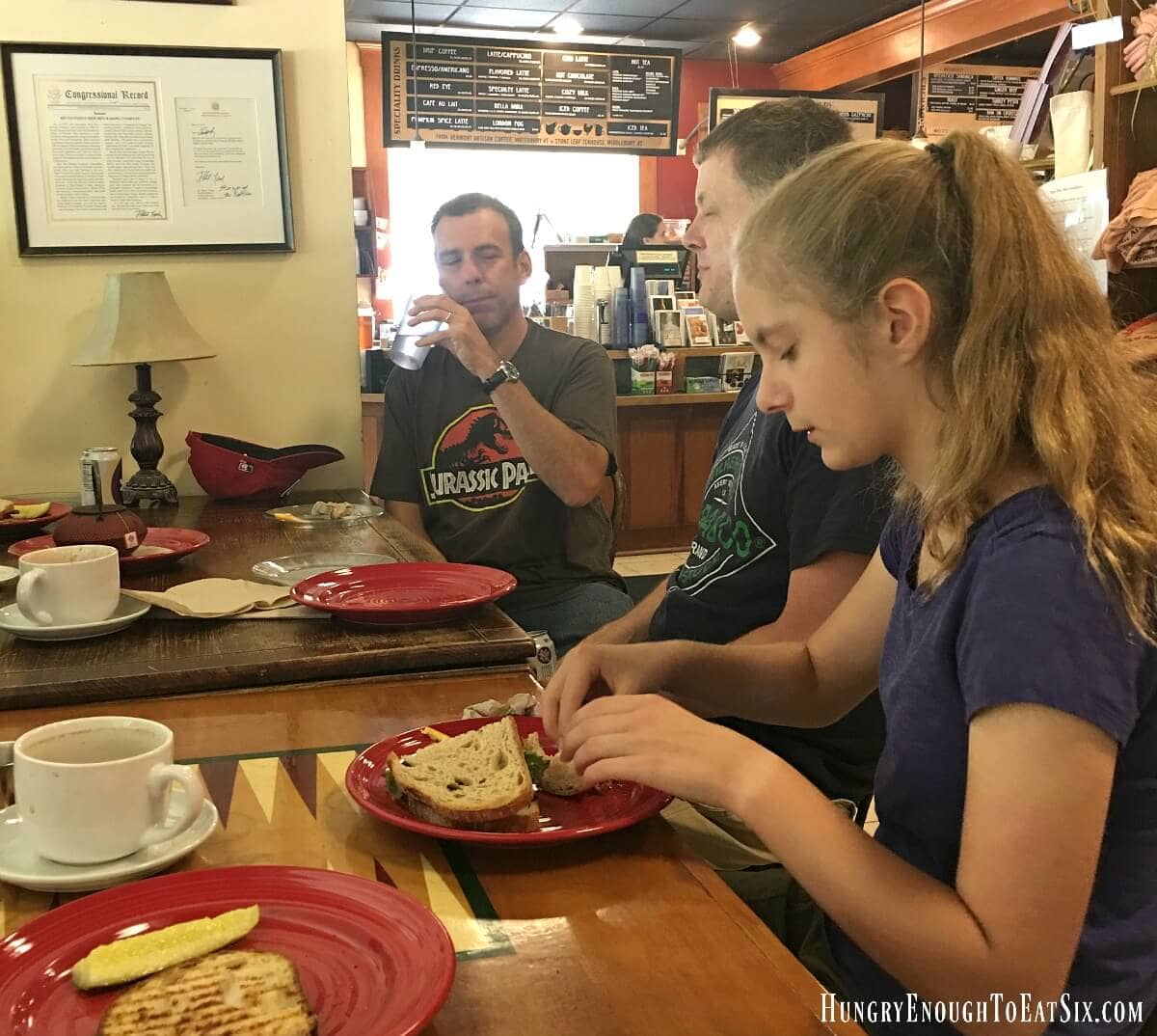 Two adults and a teen girl sitting at cafe tables eating sandwiches and having drinks.