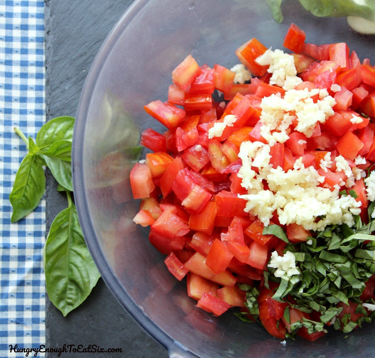 Glass bowl with separate ingredients: chopped tomato, minced garlic and basil.