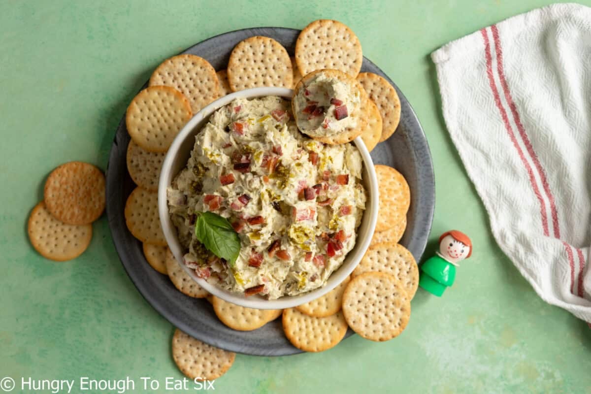 White bowl of pesto and bacon cheese spread with crackers all around it.