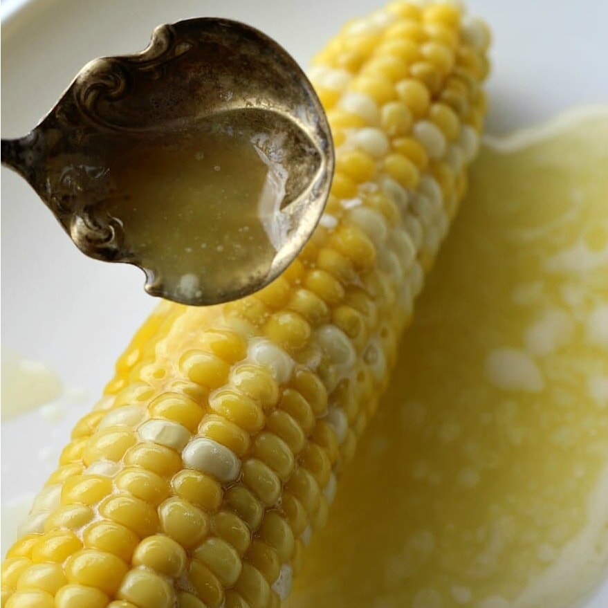 Metal ladle spooning melted butter over a cooked ear of corn on a white plate. 