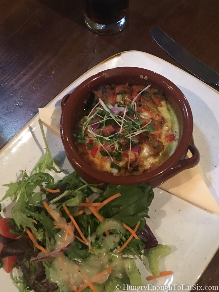 Small crock with baked vegetables and cheese on white platter.