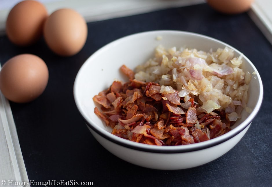 White bowl with cooked bacon and onions, whole eggs behind.