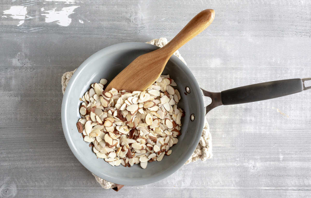 Sliced almonds in a gray skillet with a wood spoon.