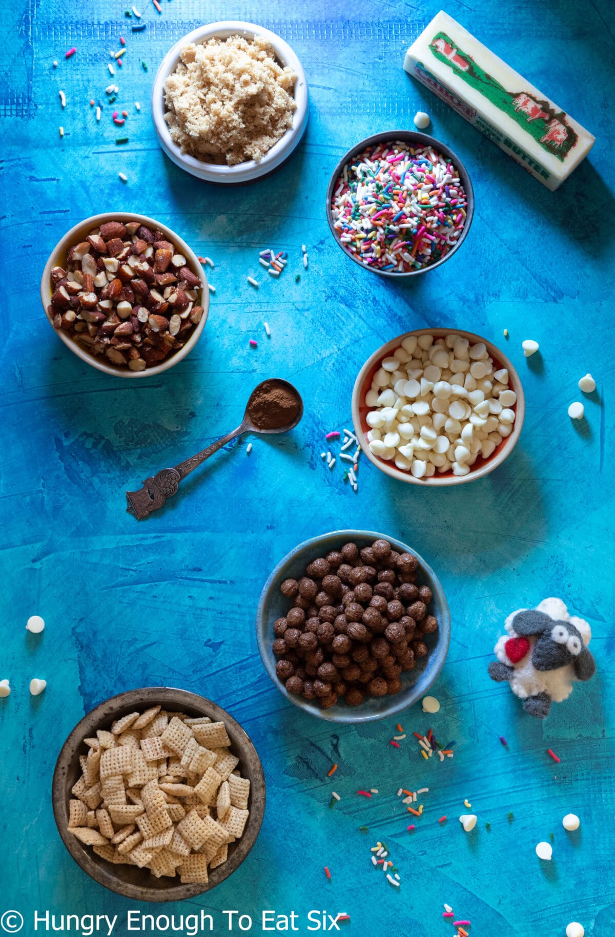 Several bowls of ingredients like Chex cereal, chocolate chips, sprinkles, Cocoa Puffs cereal, and pretzels.