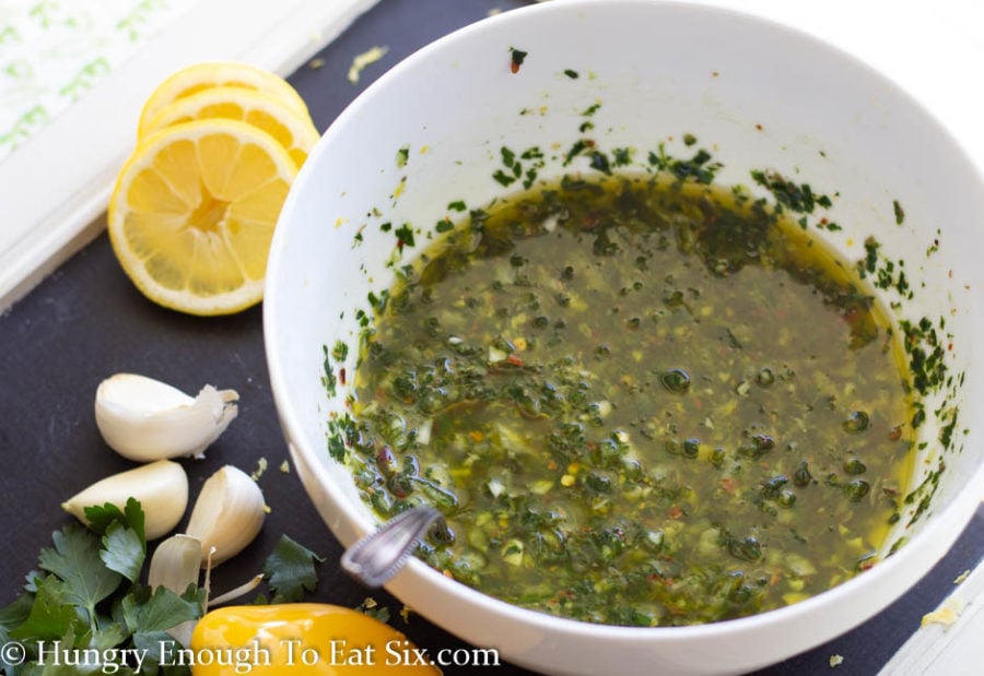 A white bowl of lemon chimichurri sauce with lemon slices and garlic cloves around it.