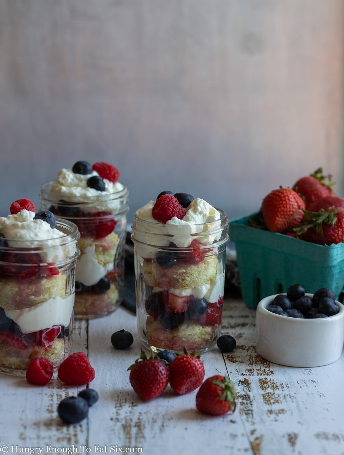 Three trifles of fruit and cream in glass jars next to containers of berries.