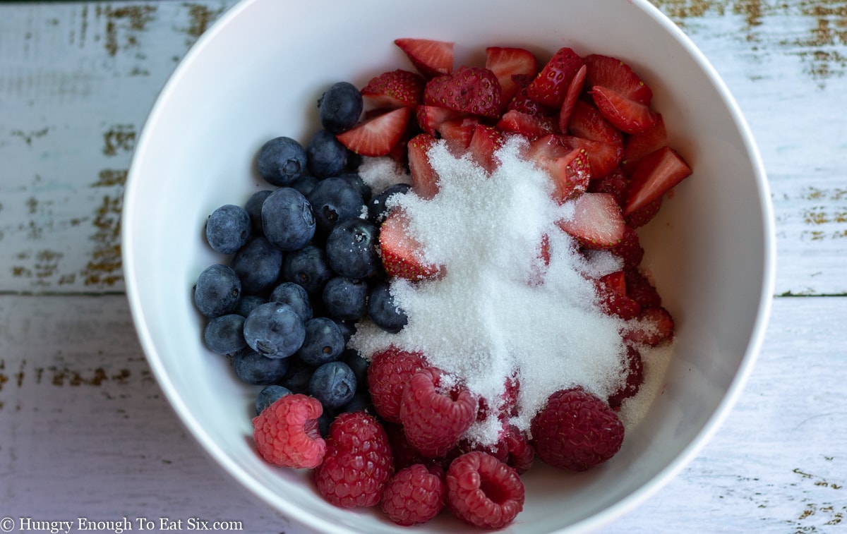 Blueberries, raspberries, strawberries and sugar in a white bowl.