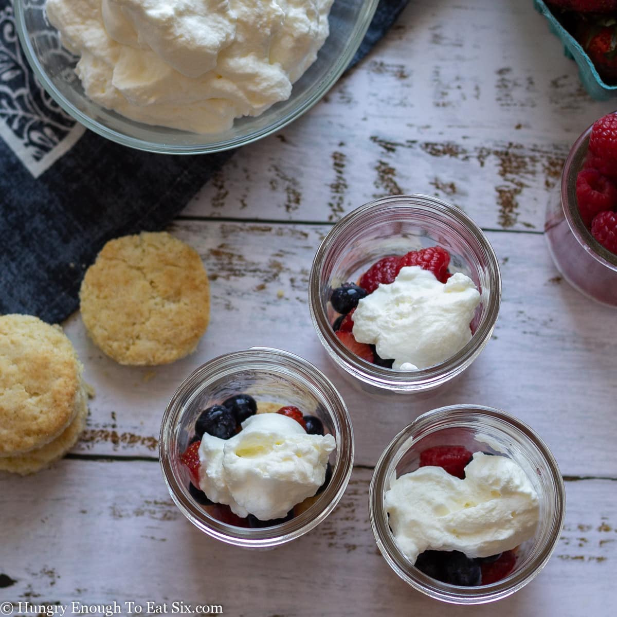 Three jars with layers of cream and berries on a white wooden surface.