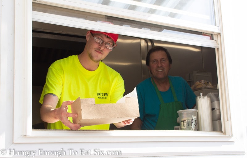 Window of Bri's Fries truck with two employees, one handing out a paper box of food. 