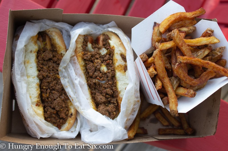 Two hot dogs in buns topped with meat sauce next to a pint of French fries. 