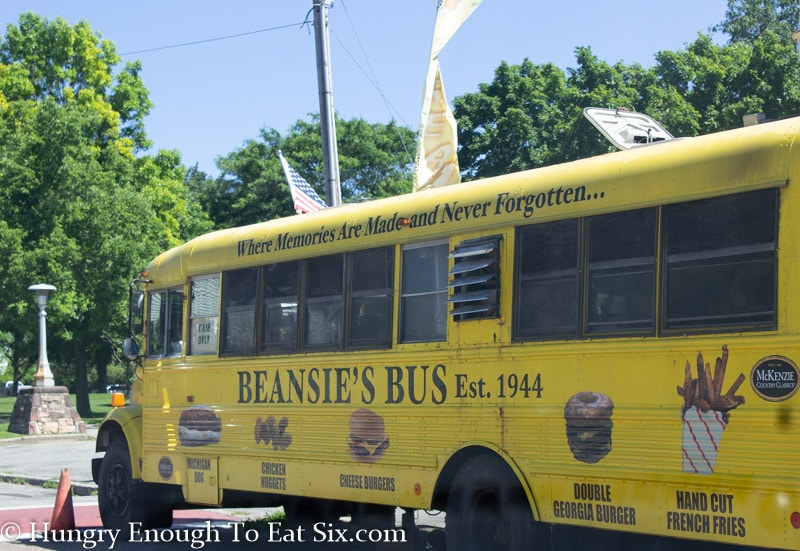 Yellow bus parked by tree-filled park, lettering on side says Beansie's Bus Est. 1944 with photos of food. 
