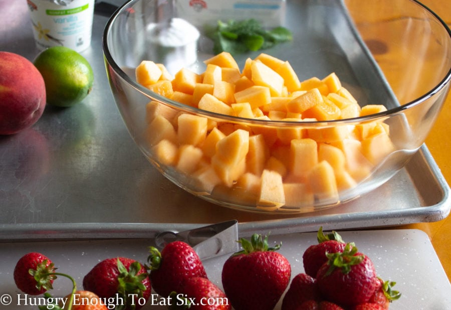 Cubed cantaloupe in a clear bowl with strawberries on a cutting board.