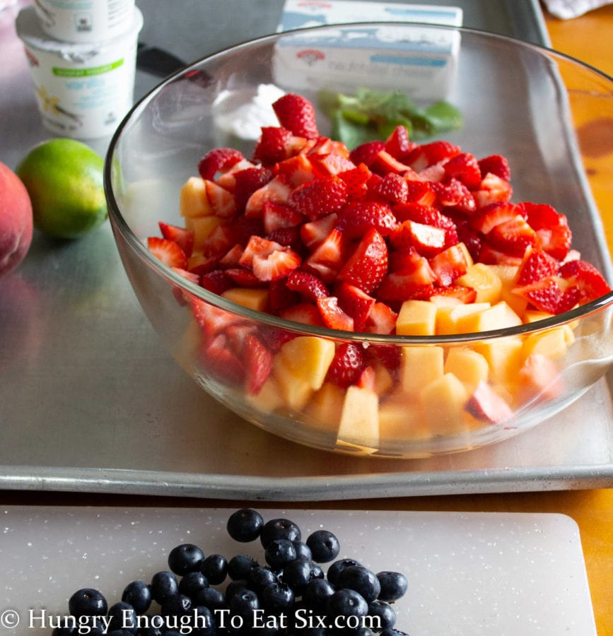 Cut strawberries and cantaloupe in a clear bowl with blueberries on a cutting board.