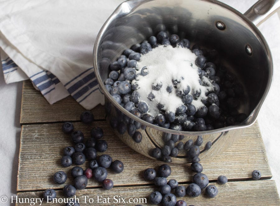Blueberries, sugar, lemon juice and water in a metal saucepan on a wood surface.