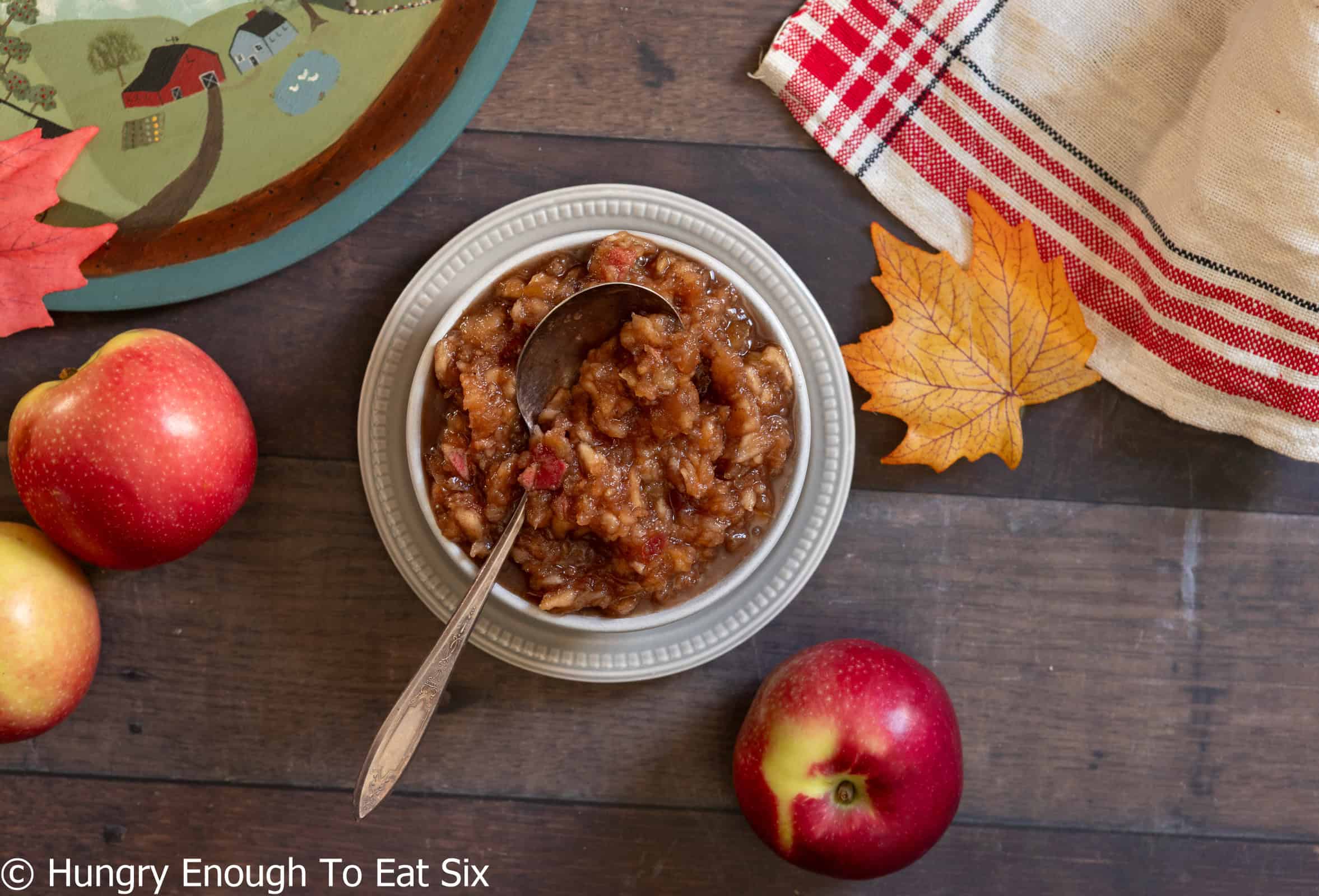 White bowl of homemade applesauce with apples and leaves nearby.