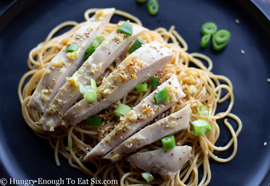 Chopped scallions and sliced chicken atop sesame noodles on a black plate.