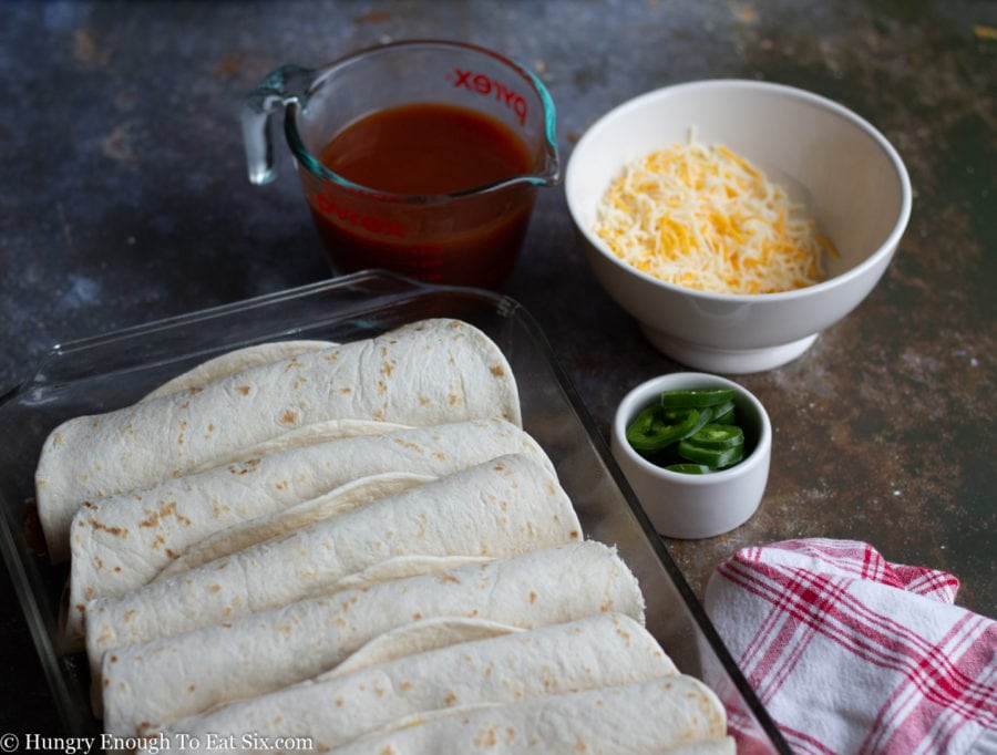 Rolled burritos in flour tortillas in a pan next to sauce and cheese.