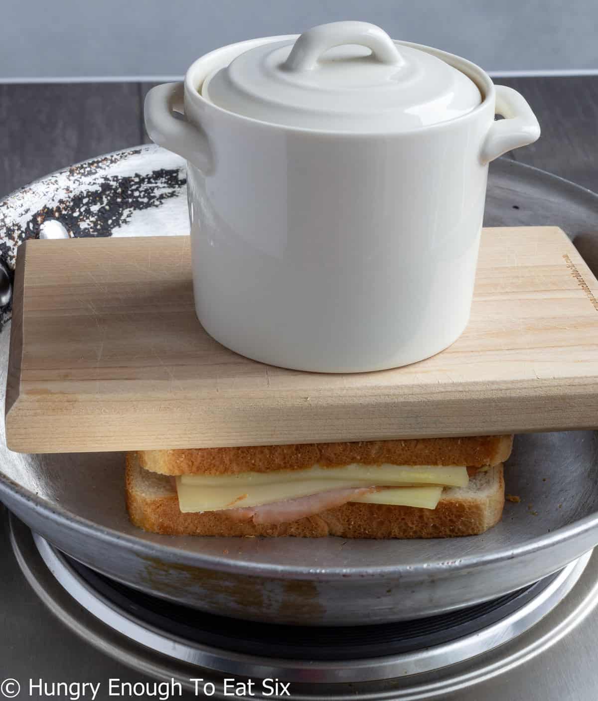 Skillet with ham and cheese sandwich under a weighted board.