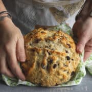 Round loaf of soda bread in hands and on green cloth.