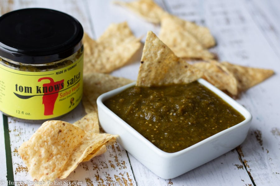 Square white dish holding salsa verde with tortilla chips all around.
