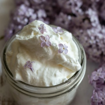 Small glass mason jar holding whipped cream with purple lilacs in background.