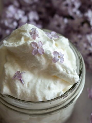 Small glass mason jar holding whipped cream with purple lilacs in background.