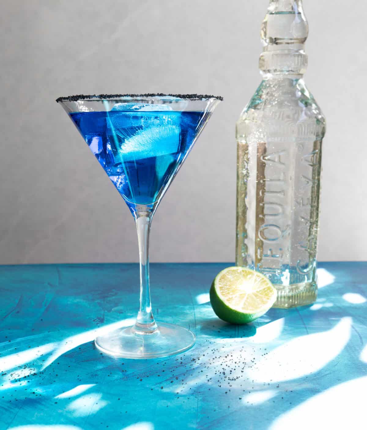 Tequila bottle, lime, and blue drink in a stemmed glass.