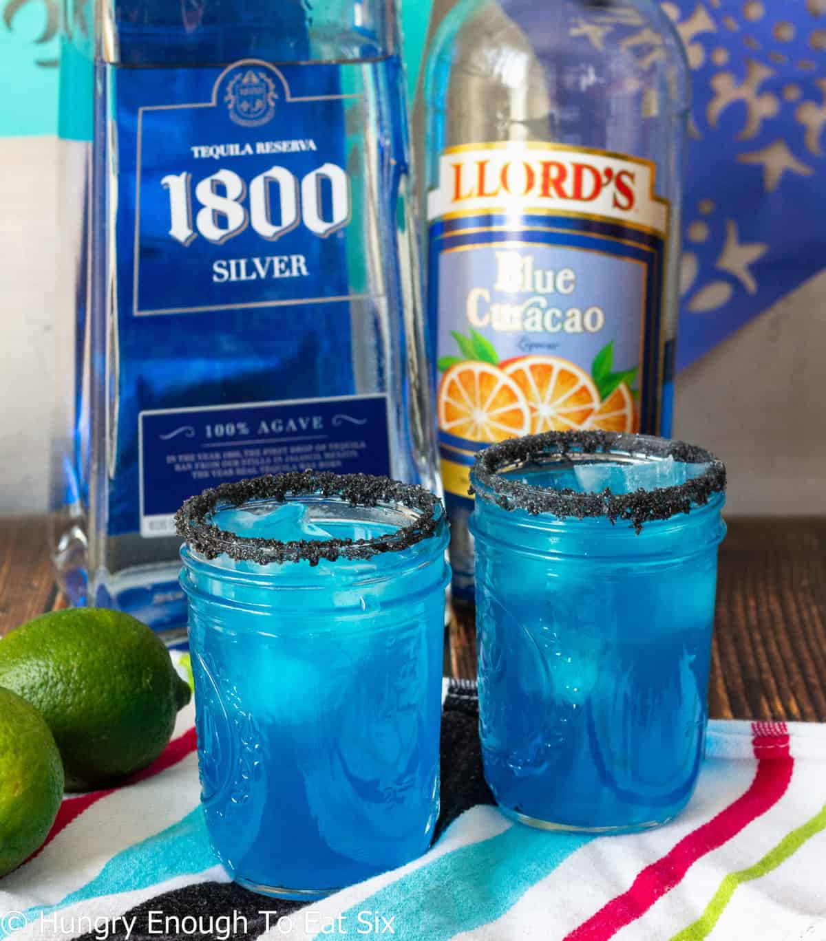 Two blue cocktails in front of bottles of tequila and blue curacao.