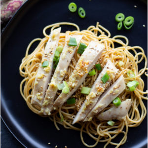Black plate with sesame noodles and sliced chicken breast, with scallions and sesame seeds.