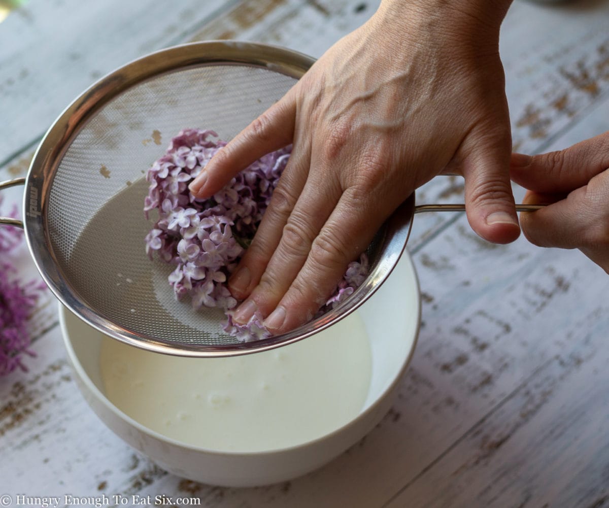 Hand pressing excess cream our of lilac sprigs in a strainer set over a bowl of cream.