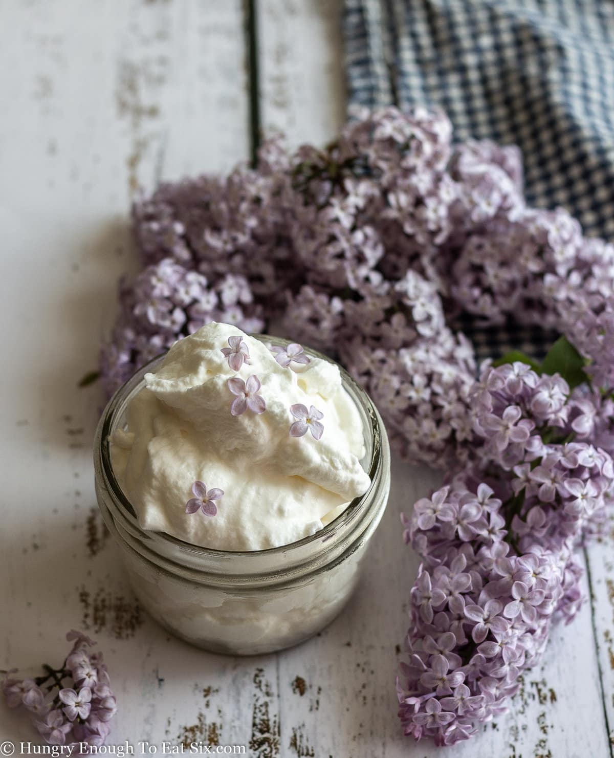 Jar of whipped cream and sprigs of lilacs laying on a white wooden surface.