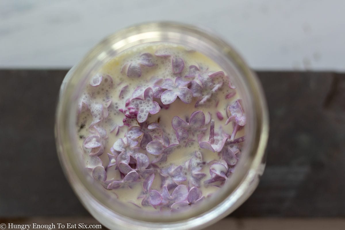 Glass jar full of cream with lilac flowers submerged in the cream.