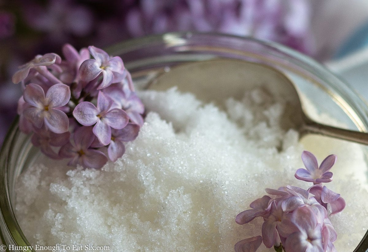White sugar sparkling in the light, and topped with purple lilac flowers and a small spoon.