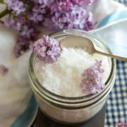 Tall image of lilac infused sugar in a glass dish surrounded by lilacs
