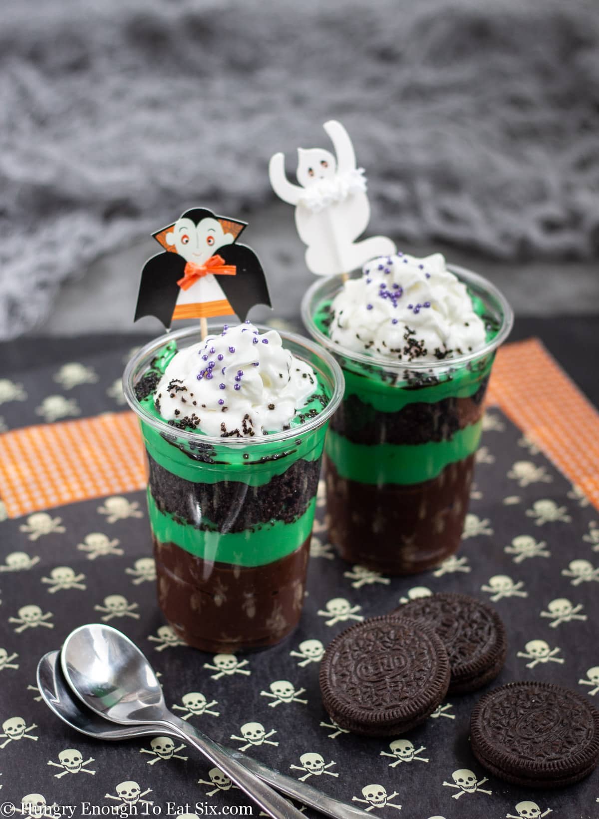 Two tall plastic cups with layered green and brown puddings and halloween decor.