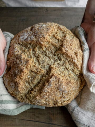 Hands holding round loaf of baked brown bread.